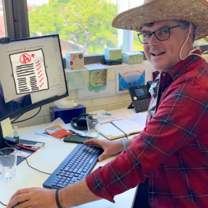 Image of CEO Leon in a red casual flanel, wearing a cowboy hat at his desk. The desk is covered in papers His computer screen is on, and a cartoon icon of an A+ graded paper is obscuring what was on his screen in the photo.