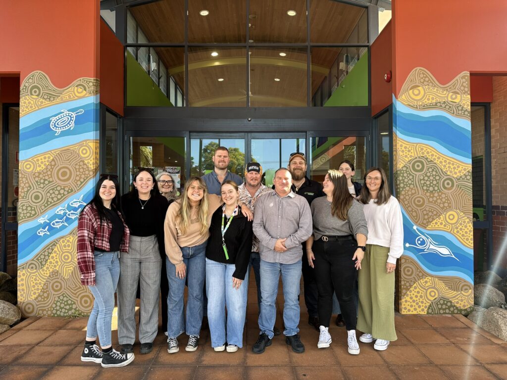 A group of thirteen people stands in front of a building entrance with glass doors. The group consists of men and women, smiling and posing closely together. The entrance features colorful, abstract artwork on both sides, depicting patterns, fish, and other elements of First Nations art. The people are dressed casually, with some wearing jeans, sweaters, and shirts. The background shows an interior space with a wooden ceiling and green walls of the Headspace youth mental health community centre.