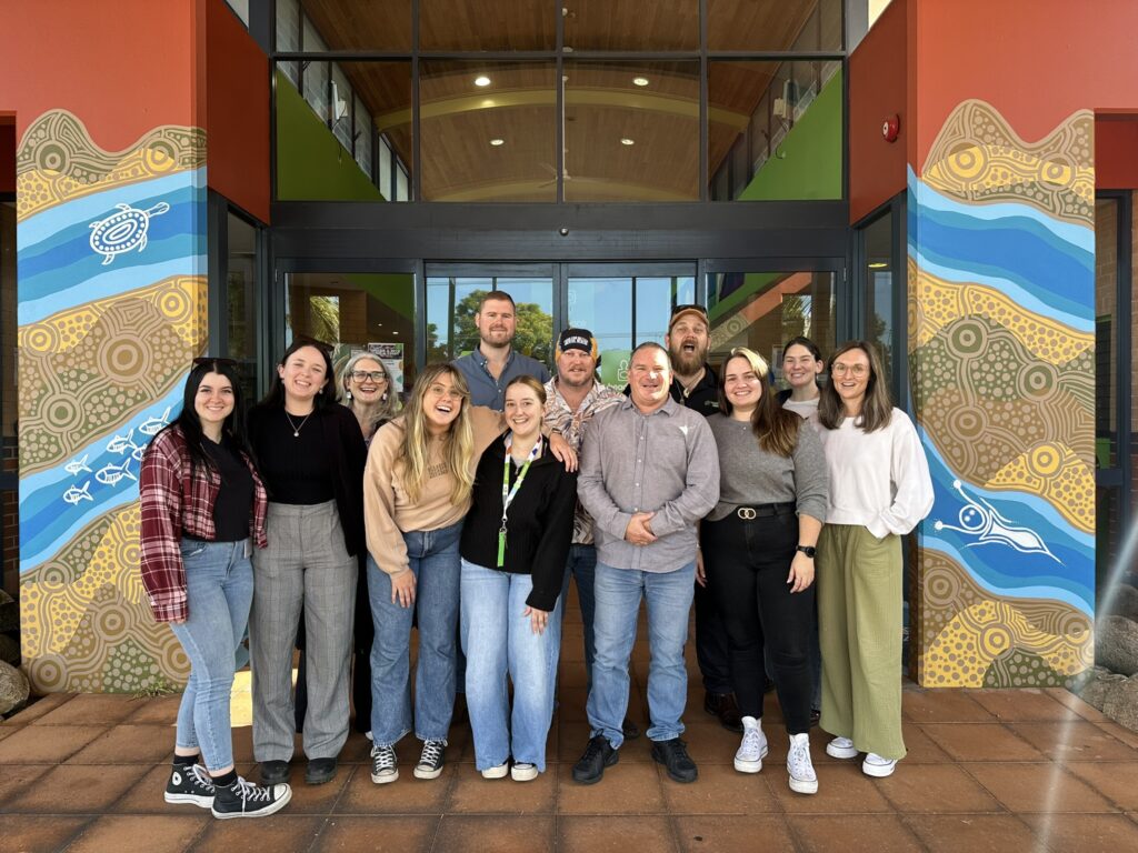 A group of thirteen people stands in front of a building entrance with glass doors. The group consists of men and women, smiling and posing closely together. The entrance features colorful, abstract artwork on both sides, depicting patterns, fish, and other elements of First Nations art. The people are dressed casually, with some wearing jeans, sweaters, and shirts. The background shows an interior space with a wooden ceiling and green walls of the Headspace youth mental health community centre.