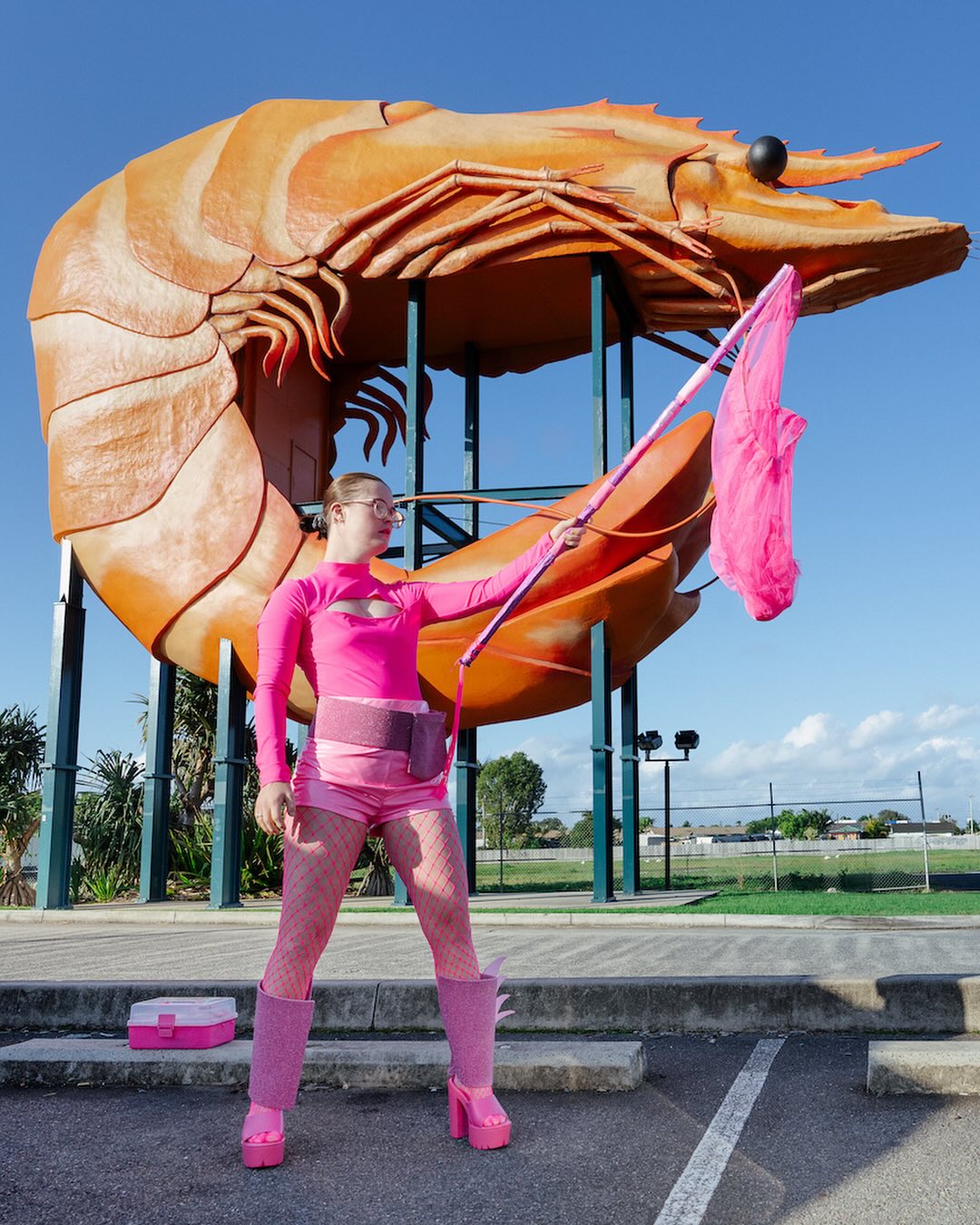 Image Description: An artist with disability wearing a superhero outfit stands in front of a giant prawn. She is holding a fishing net in her left hand with her arm raised. There are trees and a lawn in the background.