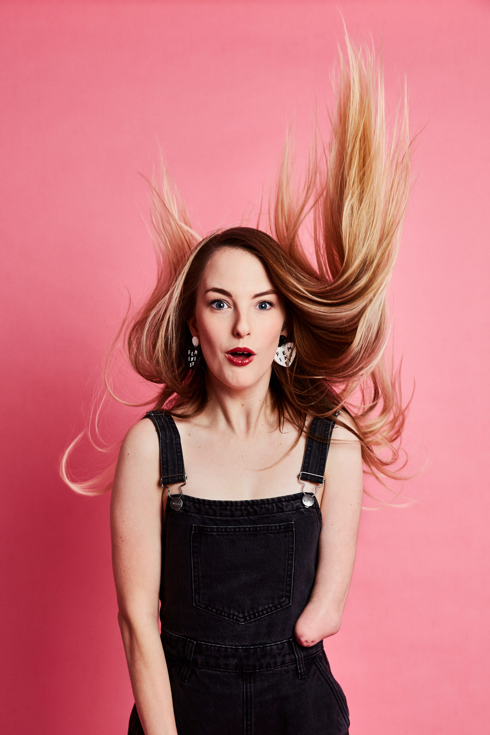 Image description: Photo of comedian Madeleine Stewart against a pink background. She is wearing black overalls and her long blonde hair is being comically blown upwards. Photo by Monica Pronk.