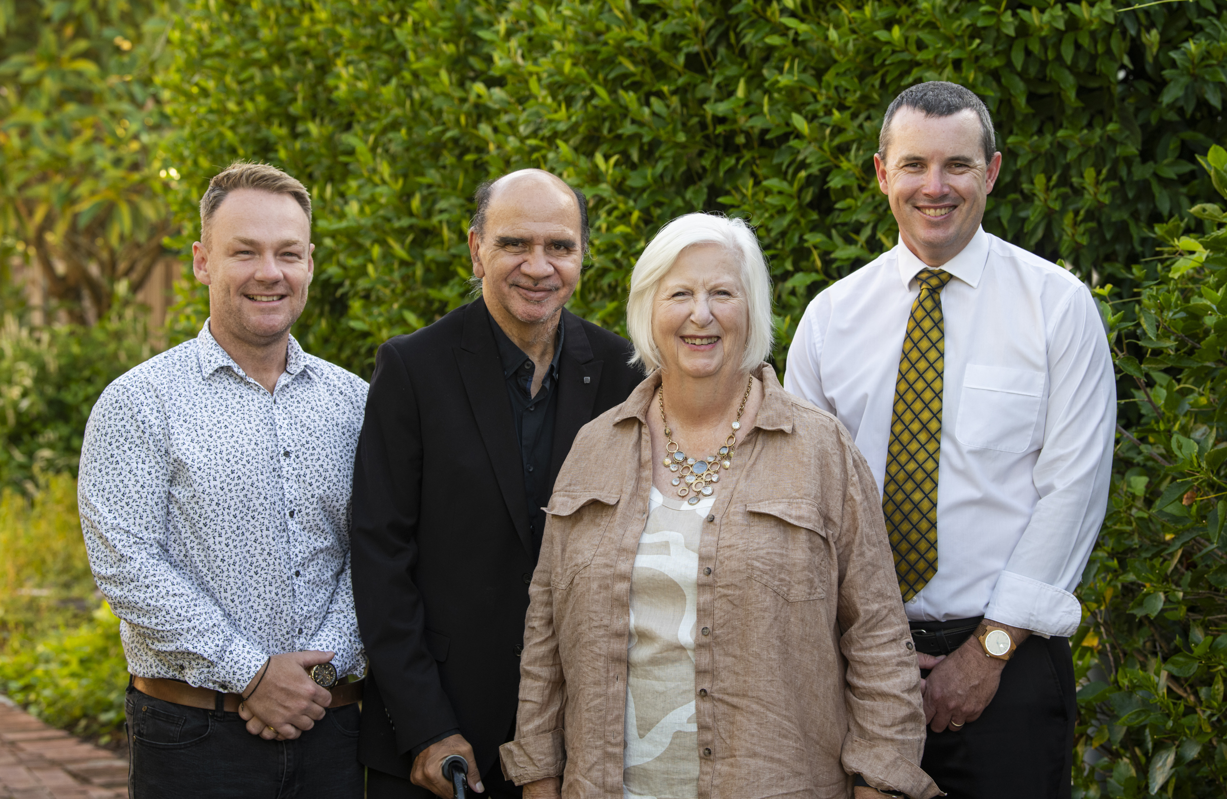 Four people standing in front of green foliage. From left to right: a man in a patterned white shirt, a man in a black suit with a walking stick, a woman in a tan jacket with white hair and statement necklace, and a man in a white shirt with yellow tie.

From left to right stand the 2025 Caringa Directors - Joshua Craig, Darren Kershaw, Vicki St Lawrence and Timothy White.